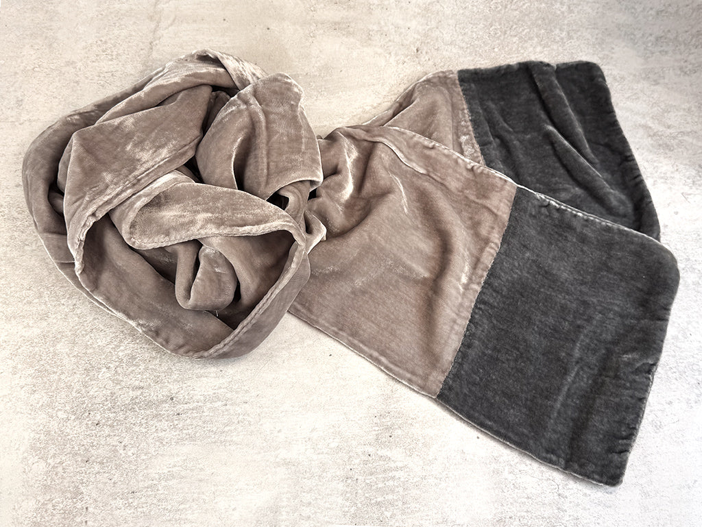 A plush velvet scarf with a colour-blocked design, featuring a taupe-brown section and a dark charcoal grey section, laid out on a textured light grey concrete surface.