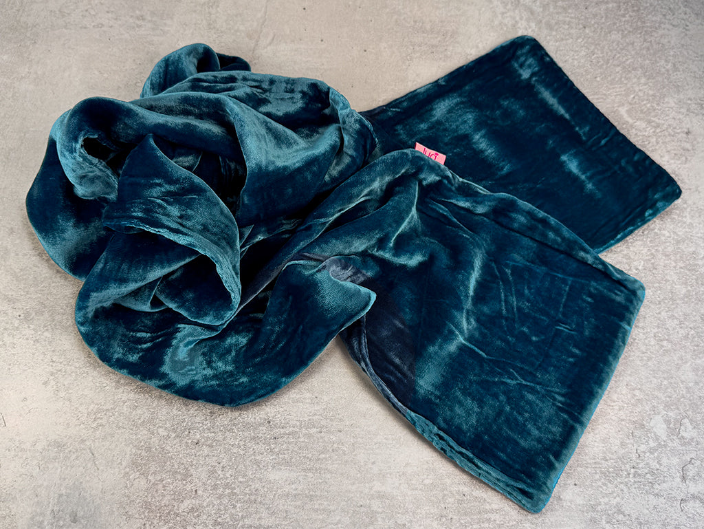 A plush velvet scarf in deep teal blue-green, folded and draped on a textured concrete surface.