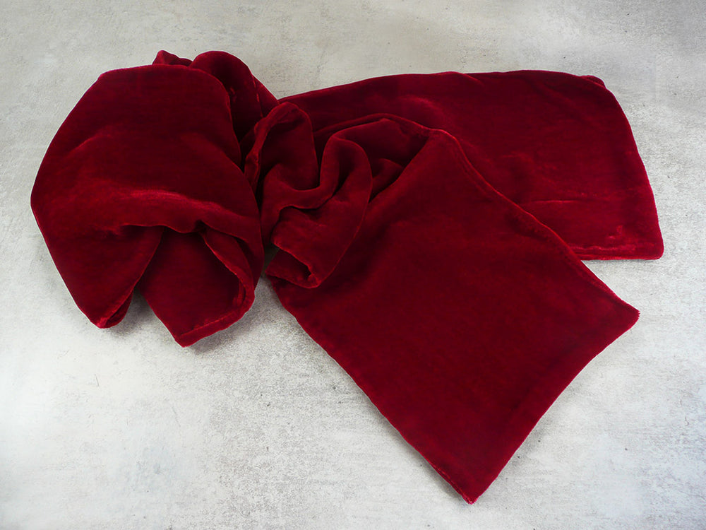 A deep red velvet scarf, styled in soft folds on a textured light grey concrete surface.