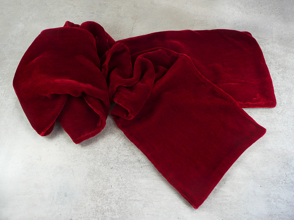 A deep red velvet scarf, styled in soft folds on a textured light grey concrete surface.